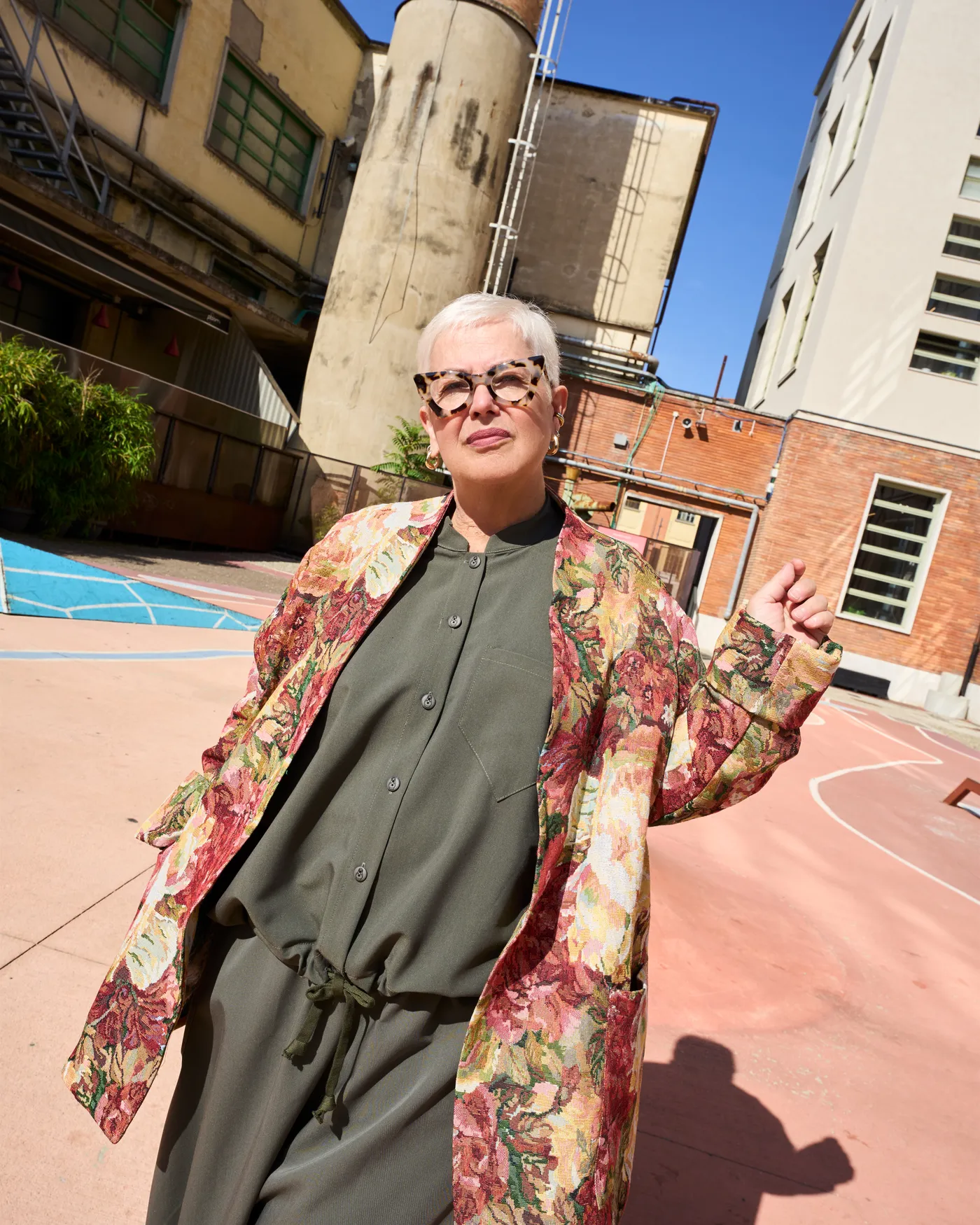 Una donna dai capelli bianchi con giacca a fiori e tuta verde oliva nel cortile della ex Manifattura Tabacchi di Firenze, con la ciminiera sullo sfondo.
