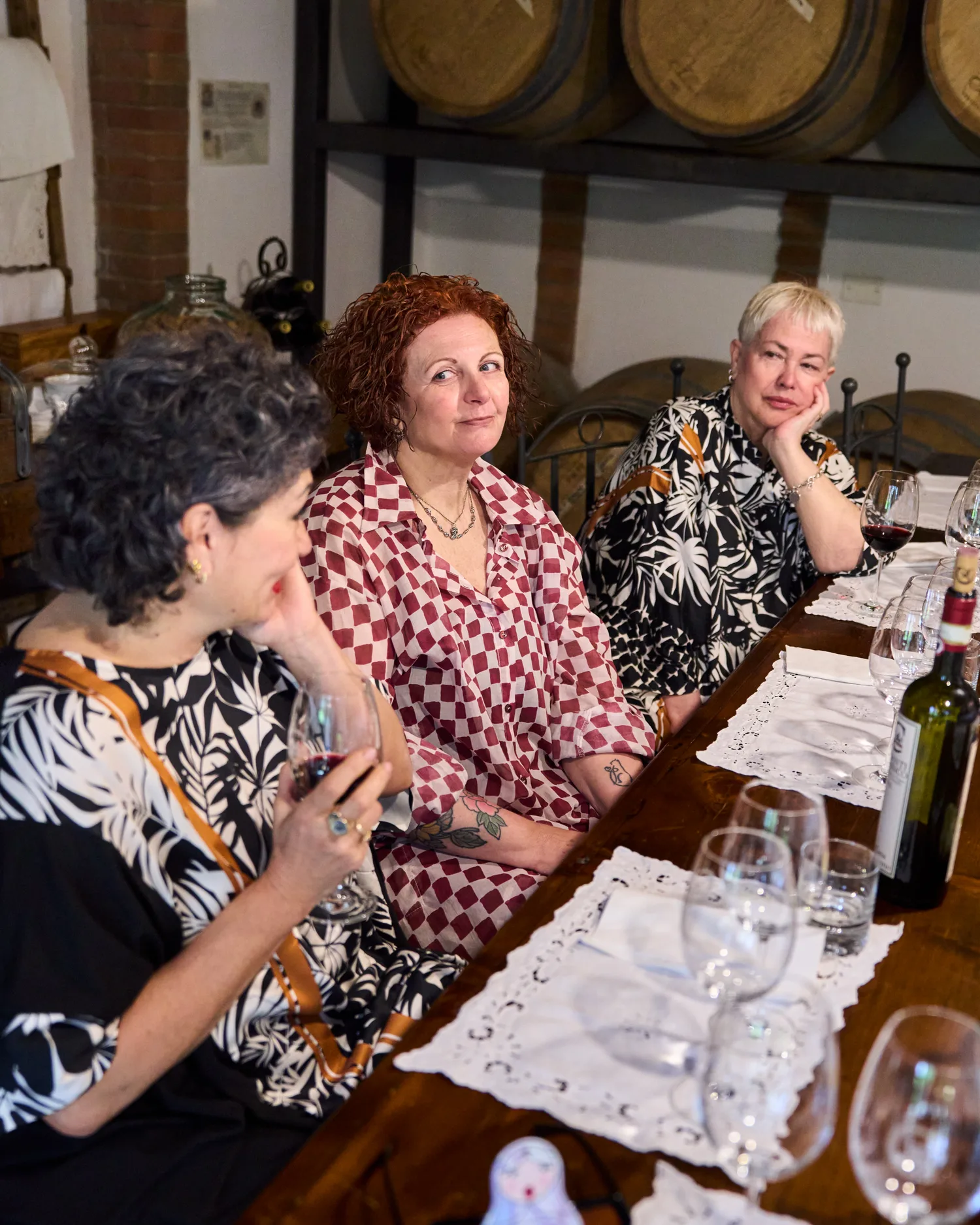 Tre donne a una tavola di degustazione in Toscana, fra legno caldo, vini rossi e stampe in bianco e nero.